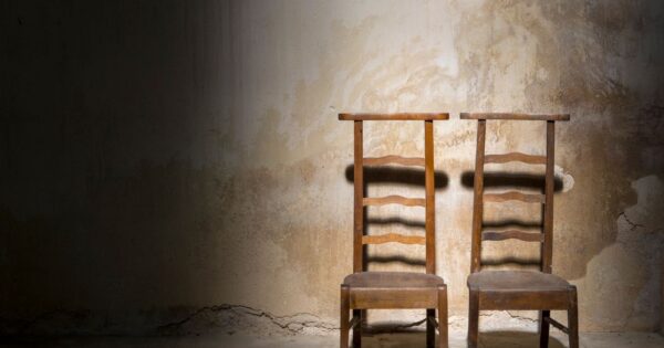 Two wooden chairs against a textured, worn wall in an empty, dimly lit room.