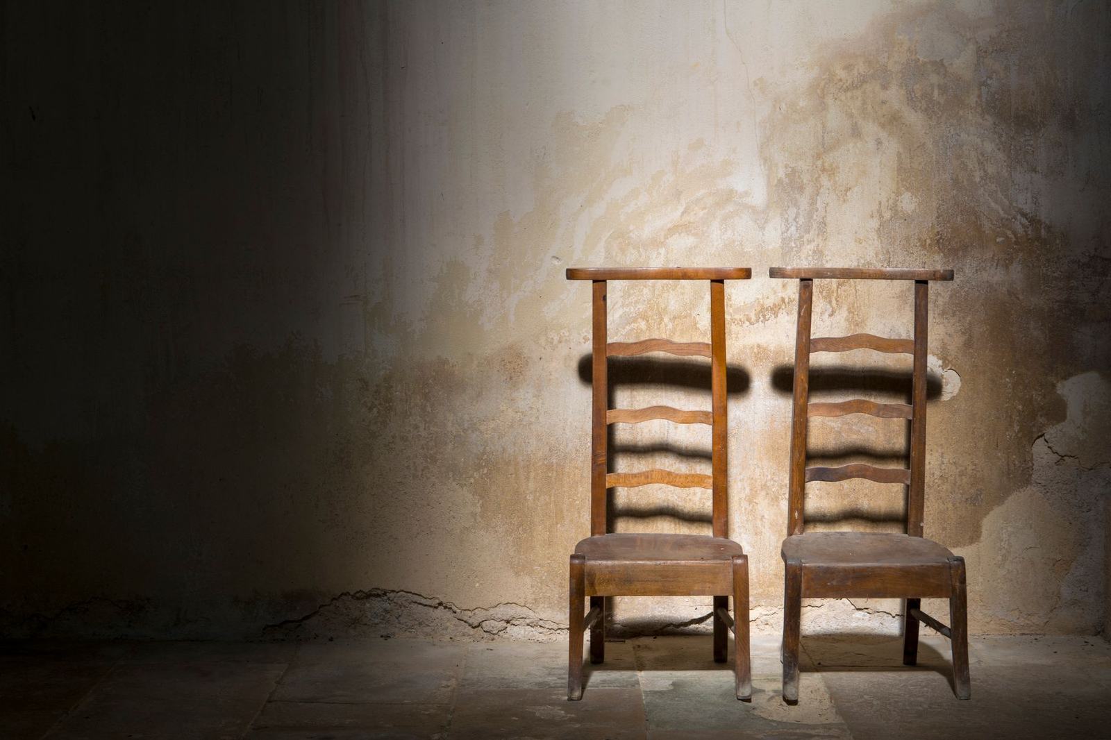 Two wooden chairs against a textured, worn wall in an empty, dimly lit room.