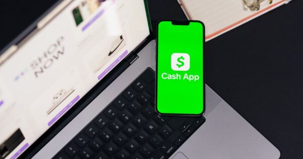 Smartphone showing Cash App screen on laptop keyboard, next to glasses and notebook.