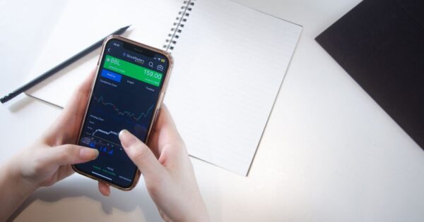 Close-up of hands using smartphone stock market app with notebook and pencil on desk.