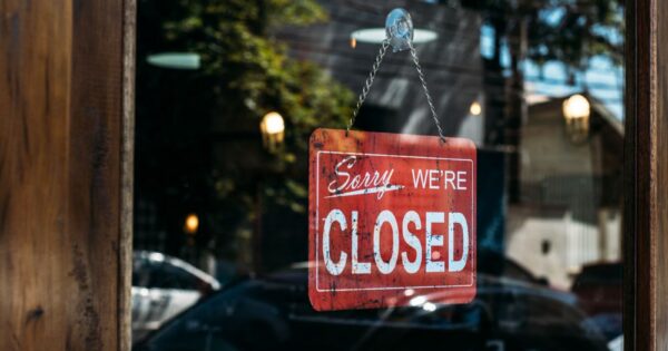 A red closed sign hangs on a glass door, reflecting cars and trees outside.