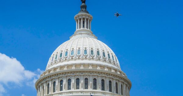 The US Capitol dome in Washington DC with a jet flying above under clear blue skies.