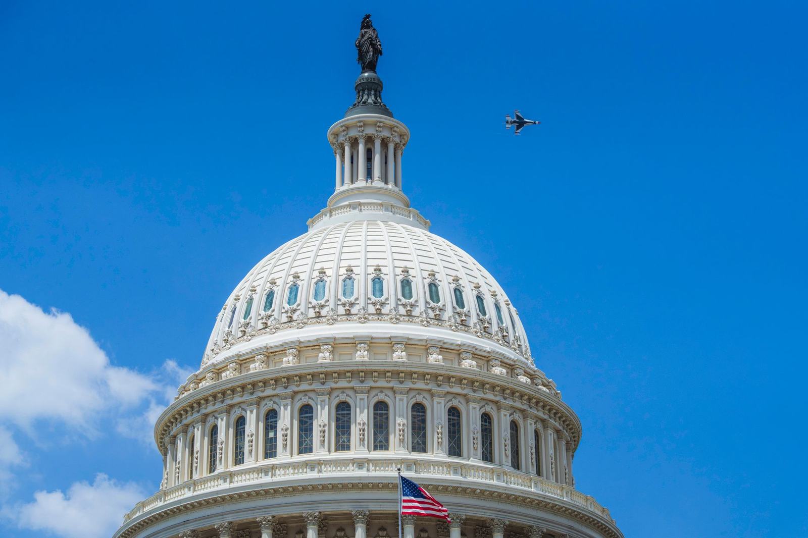 The US Capitol dome in Washington DC with a jet flying above under clear blue skies.