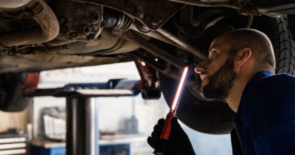 Mechanic using light to inspect vehicle undercarriage in workshop for maintenance.