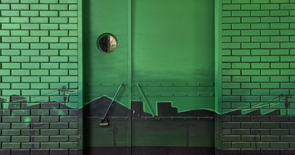 Green brick wall featuring an urban skyline mural around a door, with street art elements.