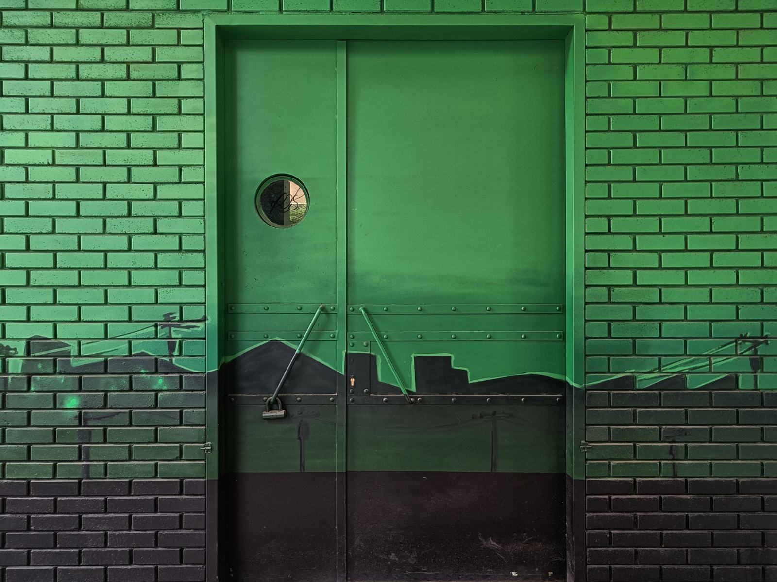 Green brick wall featuring an urban skyline mural around a door, with street art elements.