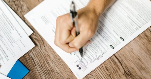 Hand writing on a W-9 tax form using a ballpoint pen on wooden desk.